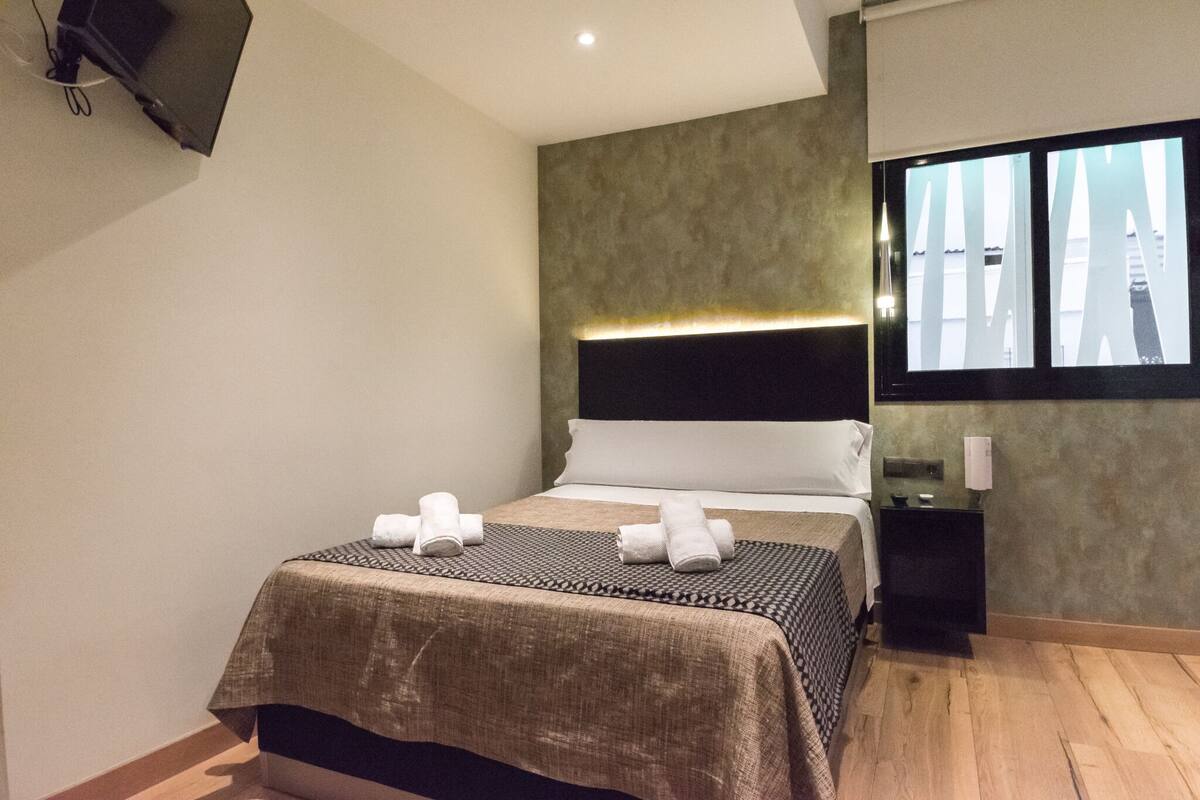 Quarto de hotel moderno com cama, mesa de cabeceira, TV na parede e janela com vista.