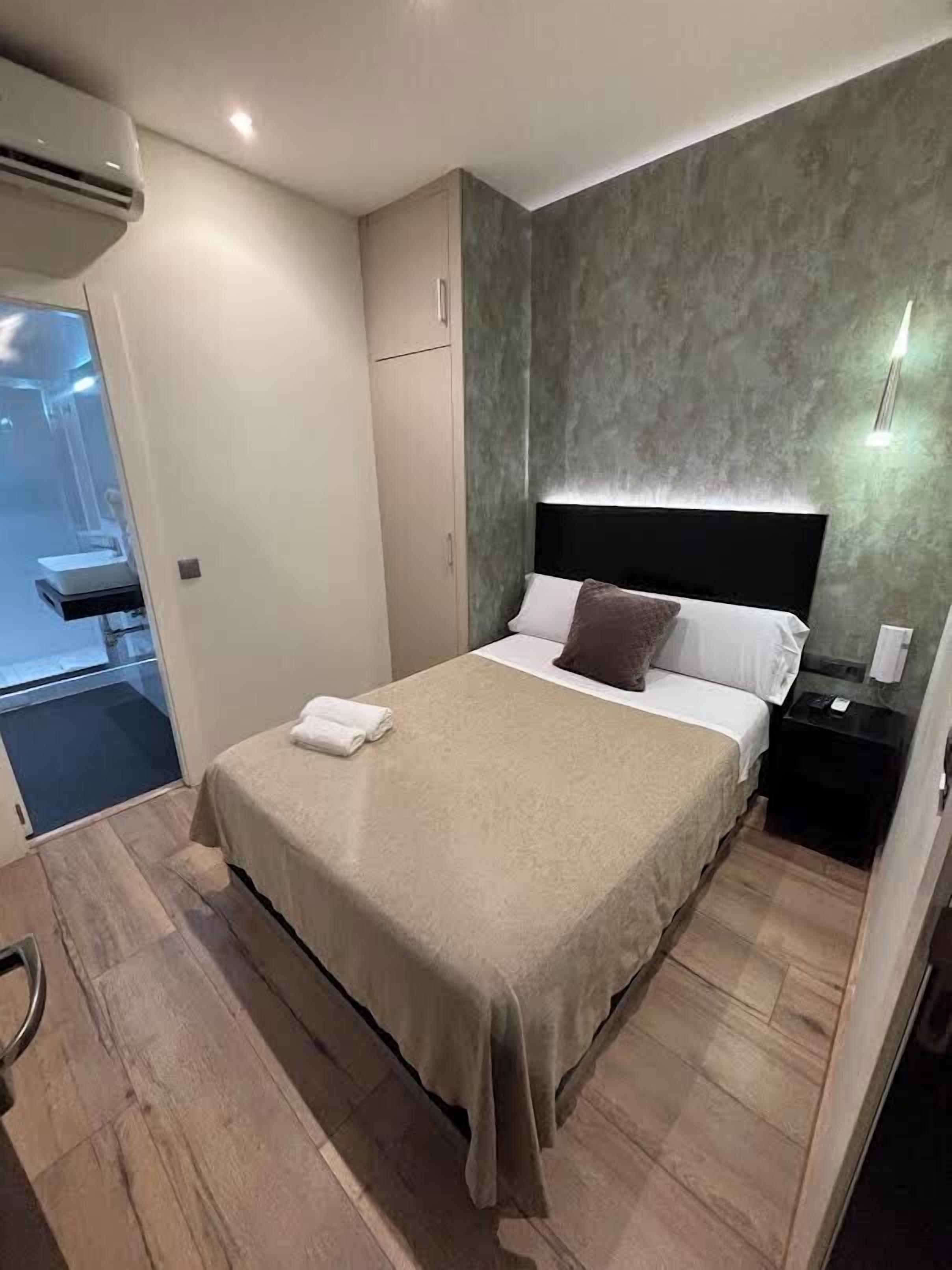 A modern hotel room with a large bed, a bedside table, a wall-mounted light, and a view of a bathroom.