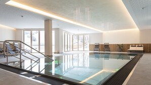 A modern indoor swimming pool with clear glass walls, a sunlit area with a reception desk, and a staircase leading to a lounge area. - Das Landerer (Ladis)