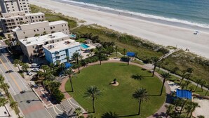 Luftaufnahme eines Strandhotels mit einer zentralen Rasenfläche, Palmen und einem Strand im Hintergrund.