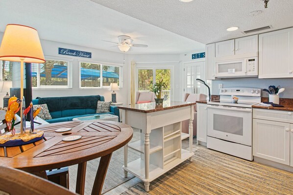 Una cocina compacta con armarios blancos y un microondas, una zona de comedor con una mesa y sillas de madera, y un sofá azul en la sala de estar.
