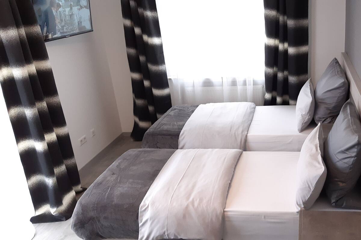 A modern hotel room with two beds, a large window with sheer curtains, and a view of the cityscape.