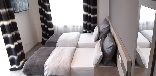 A modern hotel room with two beds, a large window with sheer curtains, and a view of the cityscape.