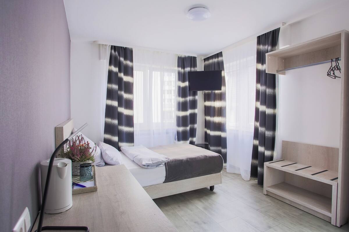 A modern hotel room with a bed, a TV, a wooden shelf, and a window with curtains.