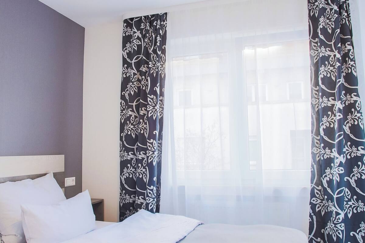A neatly made bed with white and brown linens, a wooden headboard, and a window with sheer curtains.