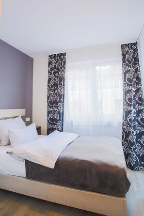 A neatly made bed with white and brown linens, a wooden headboard, and a window with sheer curtains. - City Vibes Hotel (Sindelfingen)