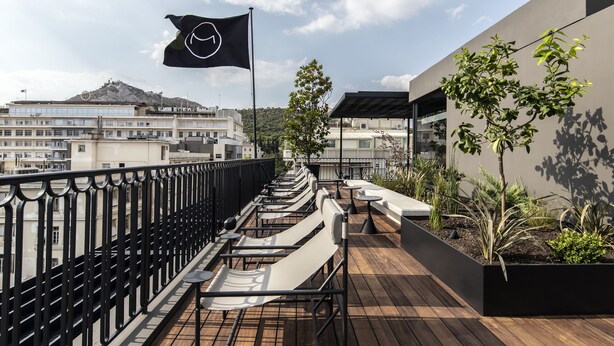 A rooftop terrace with outdoor seating, a flag, and a view of buildings and a hill in the background. - The Modernist Athens (Athens)