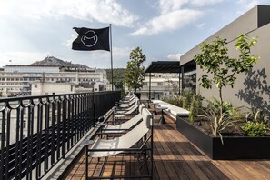 A rooftop terrace with outdoor seating, a flag, and a view of buildings and a hill in the background. - The Modernist Athens (Athens)