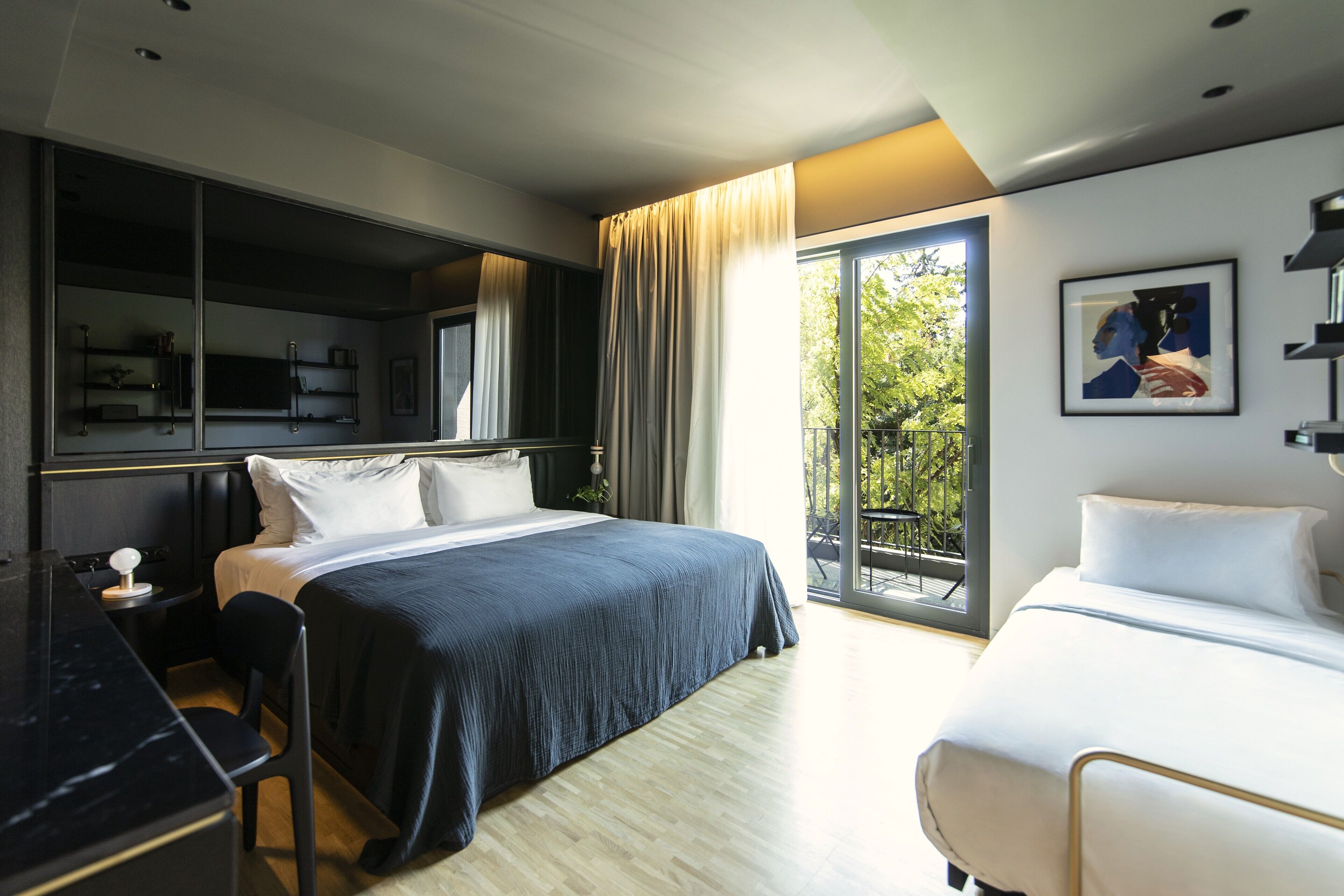 A modern hotel room with a large bed, a desk, and a balcony with a view of greenery.