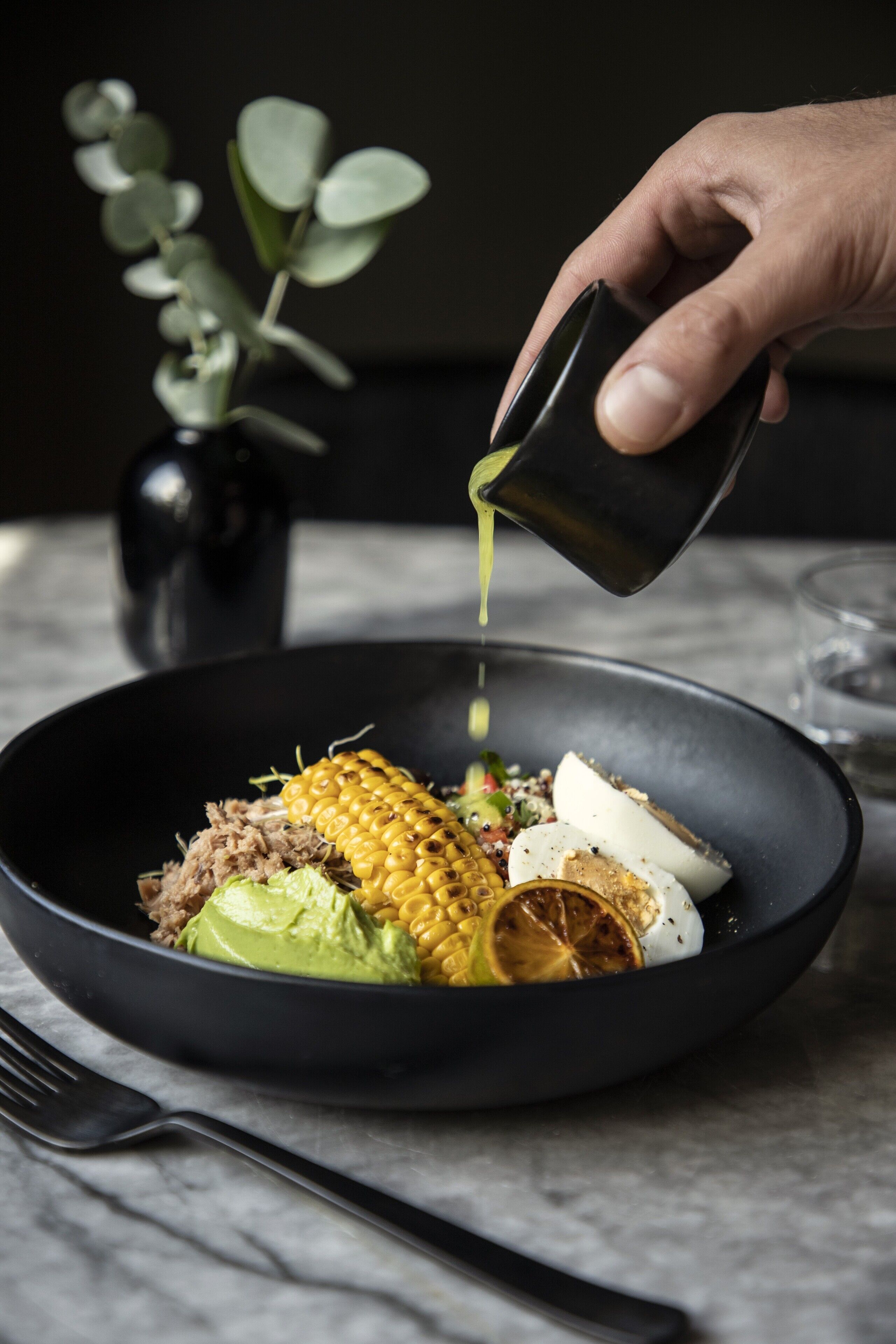 A hand pouring a green liquid into a black bowl containing food items like corn, avocado, and meat.
