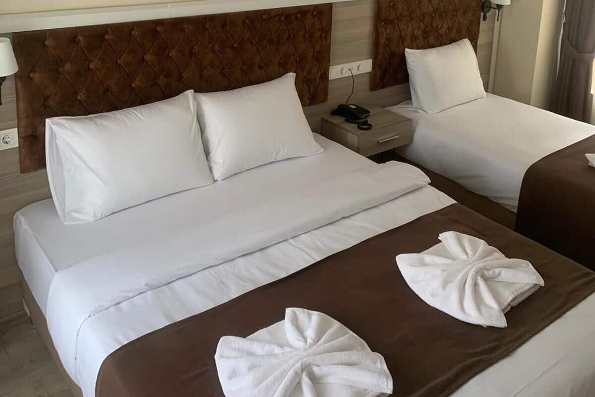 Habitación de hotel con dos camas, cada una con ropa blanca y cabeceros marrones. Hay dos mesitas de noche con lámparas y un teléfono. La habitación tiene una decoración en tonos neutros, con paredes color beige y piso de madera.