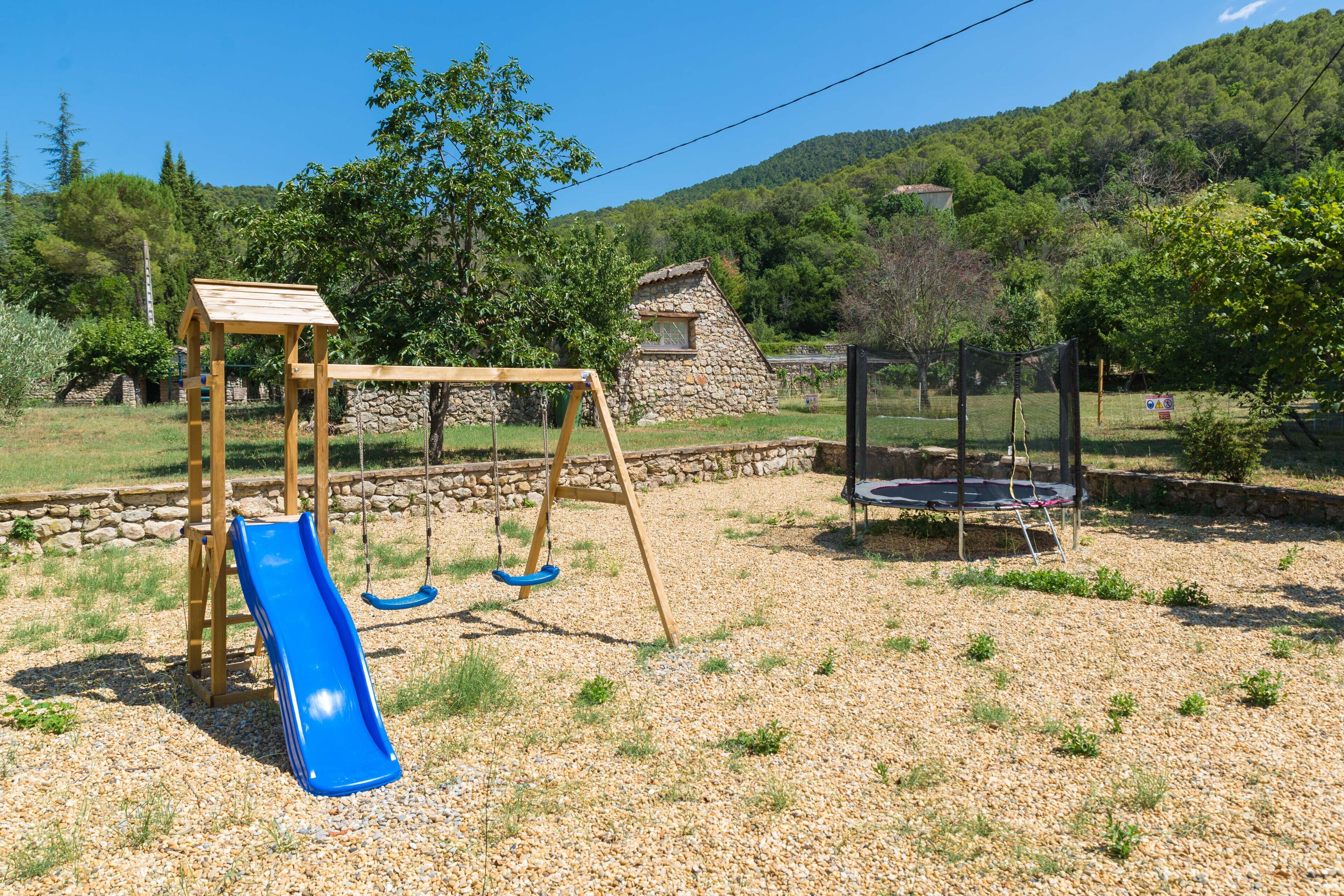Une aire de jeux avec un toboggan bleu et des balançoires, entourée d’un mur de pierre et de verdure.