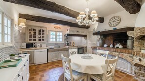 A rustic kitchen with a stone fireplace, wooden beams, and a round dining table with white chairs. - La Bastide du Riou (Bargemon)