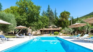A swimming pool with lounge chairs and umbrellas, surrounded by trees and a stone building. - La Bastide du Riou (Bargemon)