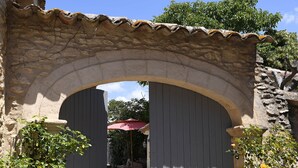 An arched stone gateway leading to a courtyard with a red umbrella and potted plants. - Bastide Des Demoiselles (Roussillon)