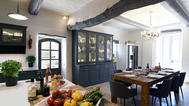 A kitchen with a wooden table set for dining, a dark blue cabinet with glass doors, and a potted plant on the counter. - Bastide Des Demoiselles (Roussillon)