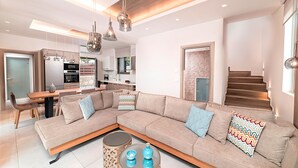 A modern living room with a sectional sofa, a dining table, and a kitchen in the background. - Minoas Maxima Villa | 2 Heated Pools | 18 guests (Georgioupoli)