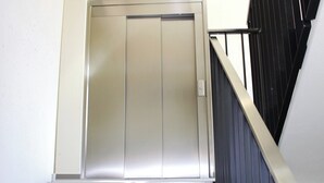 Ein moderner Aufzug mit Metalloptik, befindlich in einem Gebäude mit weißer Wand und schwarzer Geländer.