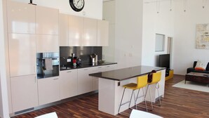 Eine moderne Küche mit weißen Schränken, einer zentralen Kochinsel mit Barhocker, einem Esstisch und einer Wohnbereiches mit Sofa und Fernseher.