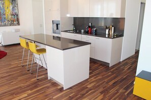 A modern kitchen with white cabinets, a black countertop, and a coffee machine. There are two yellow bar stools and a red rug on the floor. - Superb Loft In The Center Of Lausanne (Lausanne)