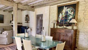 Een eetgedeelte met een glazen tafel, beige stoelen en een houten kast met daarboven een schilderij.