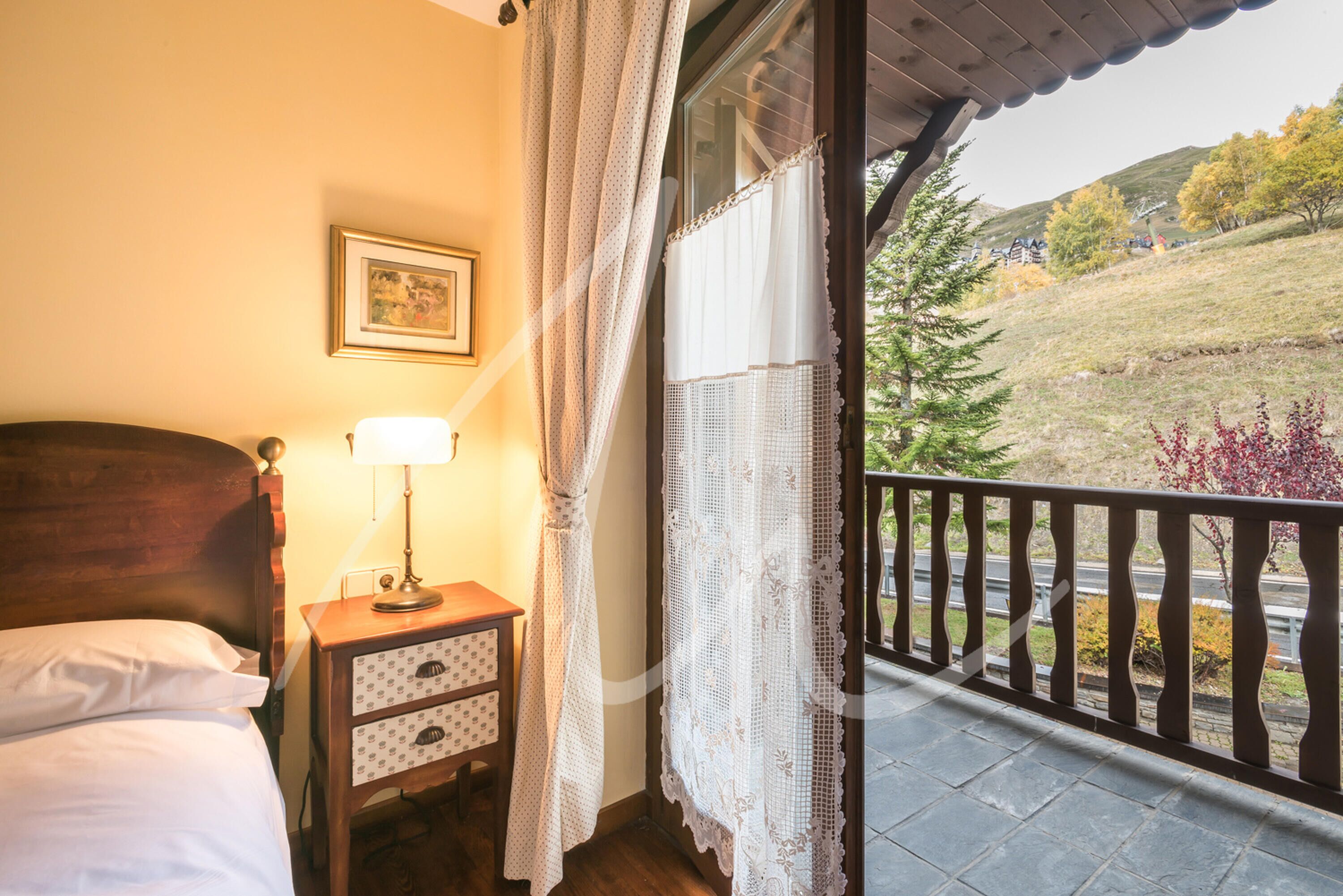Un dormitorio con cabecera de madera, una mesita de noche con lámpara, un pequeño aparador y una ventana con cortinas de encaje.
