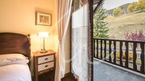 A bedroom with a wooden headboard, a bedside table with a lamp, a small drawer unit, and a window with lace curtains. - Sarrahèra (Baqueira)