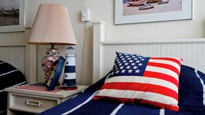 Une chambre à coucher bien rangée, avec un lit agrémenté d’un coussin orné du drapeau américain, une table de chevet avec une lampe et des objets décoratifs, et un tableau encadré accroché au mur.