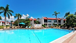 Una piscina con sillones de descanso y sombrillas, rodeada de palmeras y un edificio con tejados rojos.
