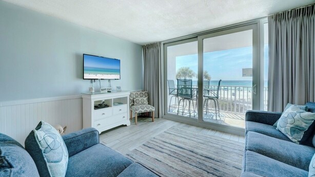 A room with a large sliding glass door leading to a balcony with a view of the sea. There is a television mounted on the wall, a white console table, and two sofas. - Edgewater Windward B-105 (Panama City Beach)