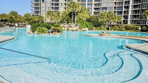 A swimming pool with clear blue water, surrounded by lush greenery and a multi-story hotel building in the background. - Edgewater Windward B-105 (Panama City Beach)
