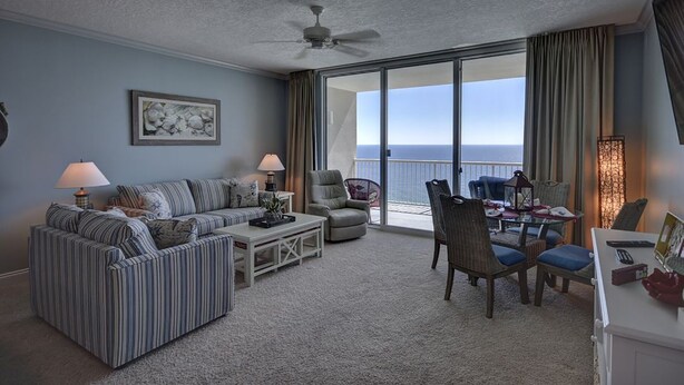 A spacious living room with a large window offering a view of the ocean, a ceiling fan, and a dining area with a table and chairs. - Palazzo 1604 (Panama City Beach)