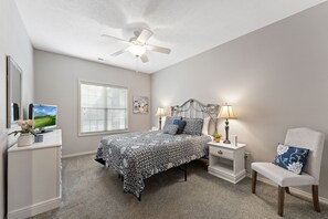 Une chambre à coucher comprenant un lit, une chaise, une table de chevet, une lampe, un ventilateur de plafond, un téléviseur et une fenêtre avec des stores.