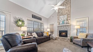 A living room with a fireplace, leather recliner, patterned armchair, and a ceiling fan.