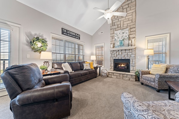 A living room with a fireplace, leather recliner, patterned armchair, and a ceiling fan.