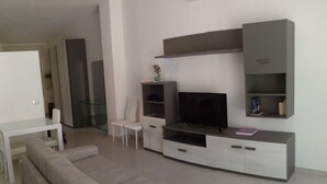 Un salón moderno con un sofá gris, un televisor sobre un mueble bajo y una mesa de comedor blanca con sillas.
