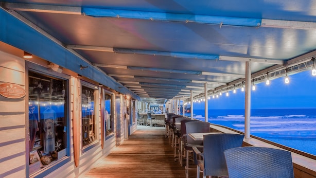 A deck with wooden flooring, tables, and chairs, overlooking the ocean.