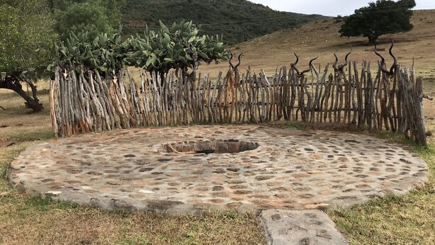 Una estructura circular de piedra con un hueco central, rodeada por una valla de madera rústica, ubicada en un campo cubierto de hierba con colinas al fondo.