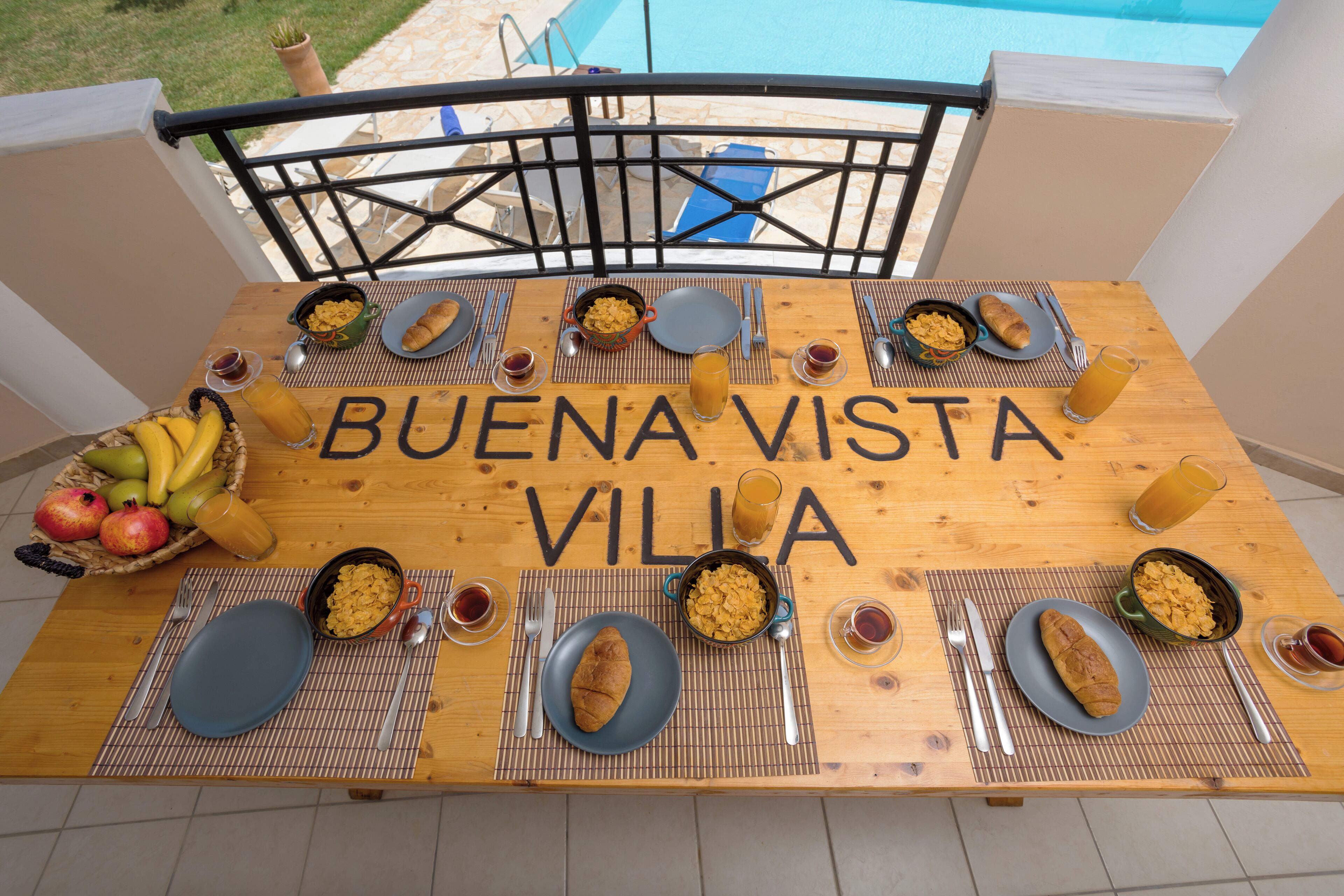 Een ontbijttafel met borden, kommen en een fruitmand, op een balkon met uitzicht op een zwembad.