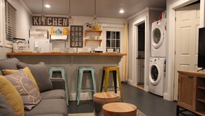A compact living space with a kitchenette, a sofa, two stools, and a washing machine.