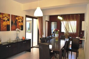Een eetruimte met een donkerhouten tafel, meerdere stoelen en een vaas met bloemen. Twee schilderijen hangen aan de muur, een groot raam met gordijnen en een hanglamp.