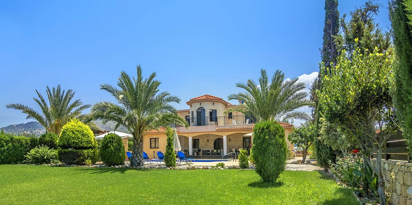 A two-story house with a balcony, surrounded by a well-maintained garden with palm trees and a swimming pool.