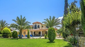 Ein zweistöckiges Haus mit Balkon, umgeben von einem gepflegten Garten mit Palmen und einem Swimmingpool.