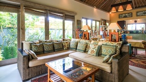 A spacious living area with a large sofa, a glass coffee table, and a dining table. There are large windows providing natural light and a view of greenery outside. - Artful 3bed3bath Villa And Bungalow in the Rice Fieldsbest Breakfast in Bali (Ubud)