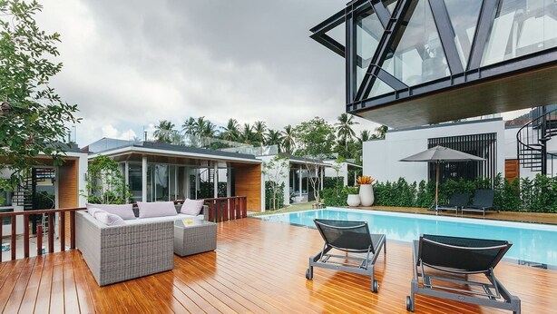 Ein moderner Poolbereich im Freien mit Holzterrasse, einer Lounge mit Korbmöbeln und einer gläsernen Struktur.