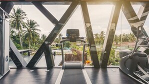 Ein Fitnessraum mit Laufband, Crosstrainer und Heimtrainer, mit einem großen Fenster und Blick auf Palmen und eine Straße.