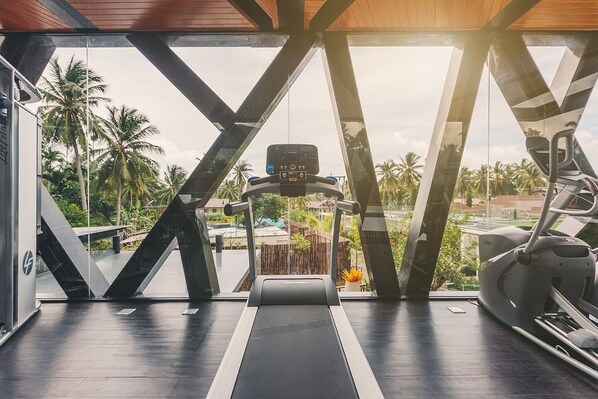 Gimnasio con cinta de correr, elíptica y bicicleta estática, con un amplio ventanal que ofrece vistas a palmeras y una carretera.
