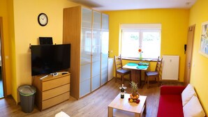 A compact living space with a television, wooden furniture, a dining area with a table and chairs, and a window with a view. - Havelapartments Gatow (Berlin)