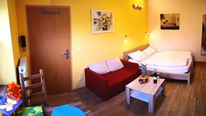 A hotel room with a wooden door, a red sofa, a bed with white bedding, a small table with a pineapple and wine bottle, and a painting on the wall. - Havelapartments Gatow (Berlin)