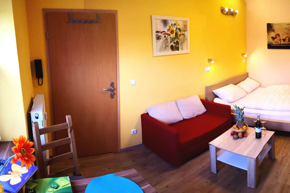 Une chambre d’hôtel avec une porte en bois, un canapé rouge, un lit avec une literie blanche, une petite table sur laquelle se trouvent un ananas et une bouteille de vin, et un tableau accroché au mur.